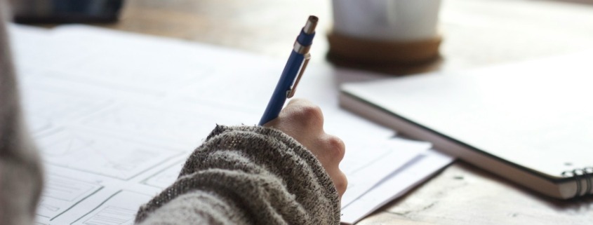 person writing on brown wooden table near white ceramic mug